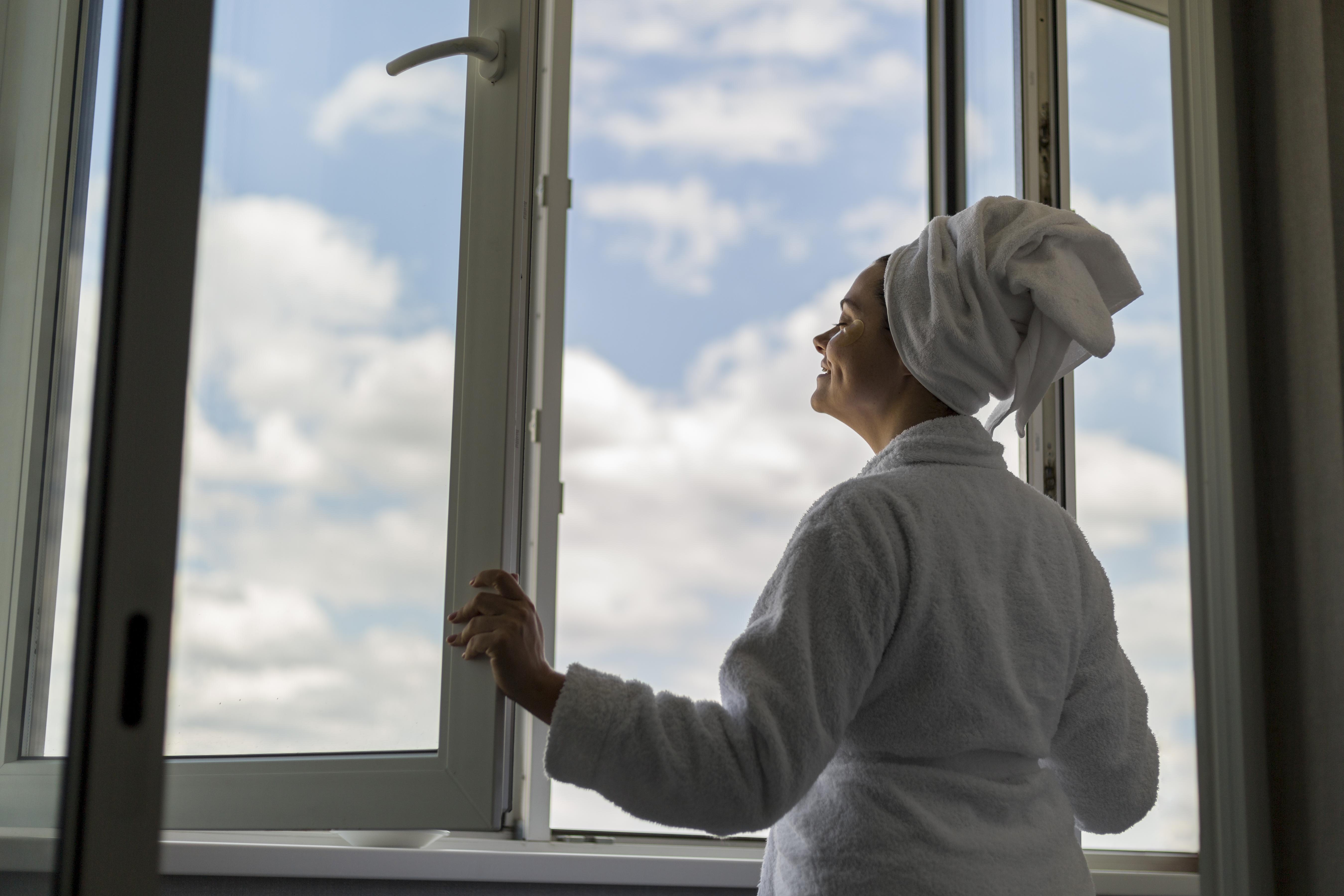 Frau im Bademantel &ouml;ffnet ein Fenster mit Dreifachverglasung und blickt in den Himmel mit Wolken.