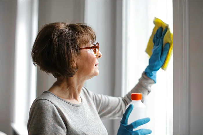 &Auml;ltere Frau reinigt ein Fenster mit einem Tuch und h&auml;lt eine Flasche Glasreiniger in der Hand.