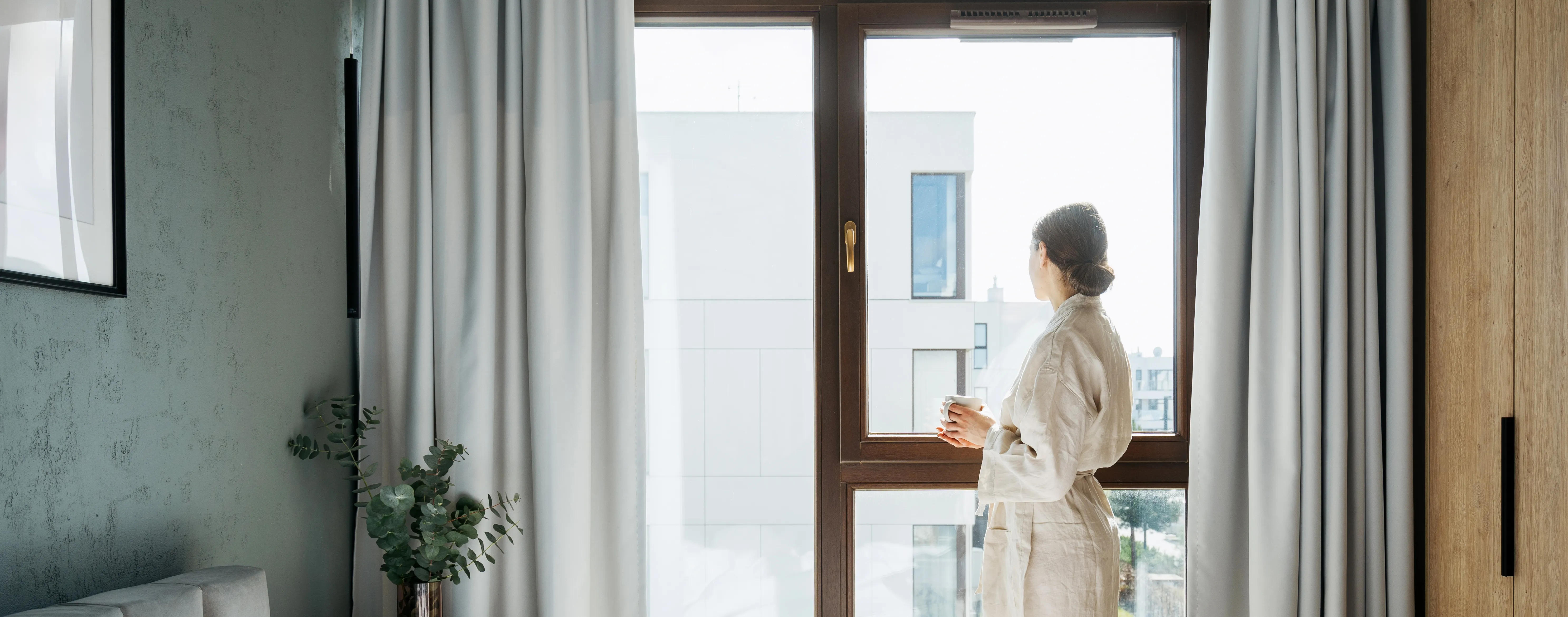 Frau steht am Fenster mit einer Tasse in der Hand und blickt durch ein gro&szlig;es Wohnungsfenster nach drau&szlig;en.