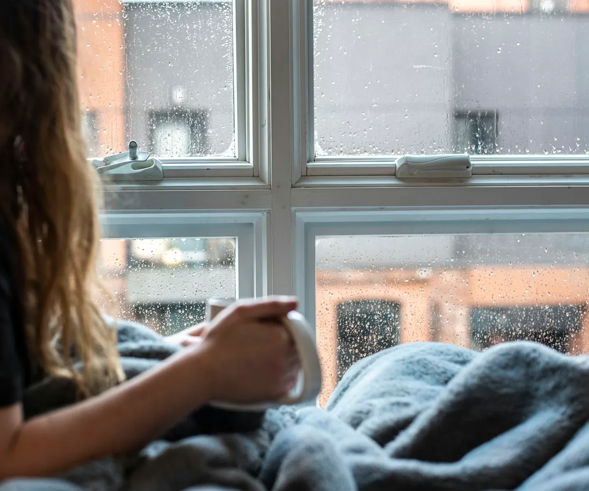 Person sitzt mit einer Tasse am Fenster, auf der Scheibe sind Feuchtigkeitstropfen sichtbar &ndash; Beispiel f&uuml;r Kondensation oder eine undichte Isolierverglasung.
