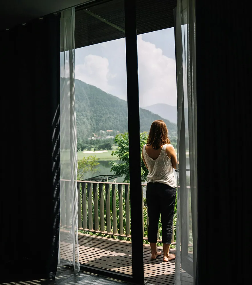 Frau auf einem Balkon vor einer geöffneten verglasten Balkontür mit Blick auf die Berge