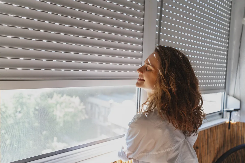 Frau am Fenster mit heruntergelassenem Rollladen &ndash; effektiver Sonnenschutz zur Reduzierung von Hitze im Innenraum.