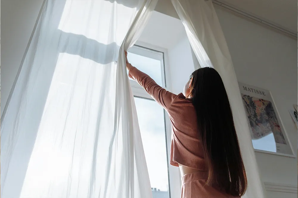 Frau öffnet Vorhänge am Fenster für richtiges Lüften – Maßnahme zur natürlichen Kühlung von Räumen im Sommer.