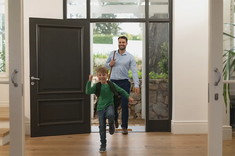 Vater und Kind betreten ein Haus durch gro&szlig;e verglaste T&uuml;r &ndash; Beispiel f&uuml;r R&auml;ume mit viel Glasfl&auml;che, die sich im Sommer schneller aufheizen.