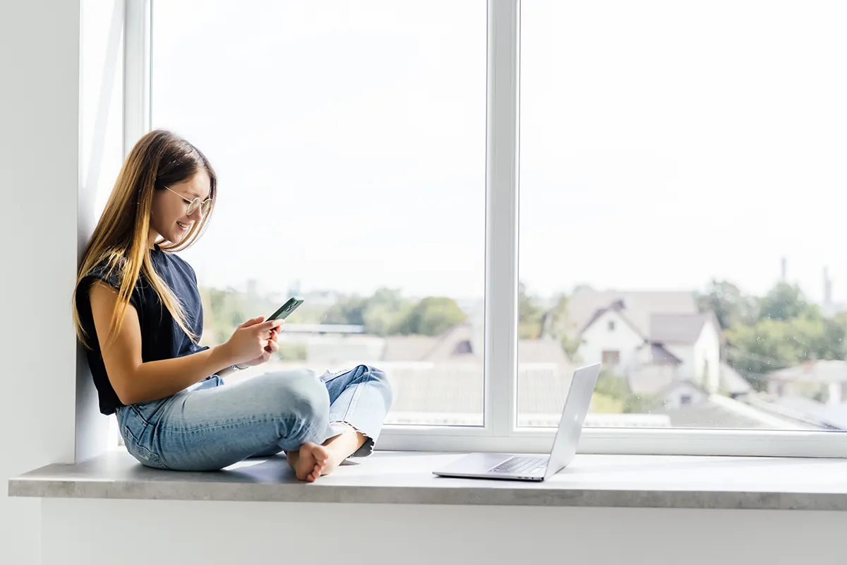 Junge Frau sitzt auf der Fensterbank mit Smartphone – modernes, helles Interieur als Symbol für komfortables Wohnen mit energieeffizienten Fenstern.